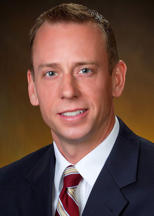 Todd Hingson, a man in a suit and tie, smiles at the camera. He has short brown hair and is wearing a dark jacket with a white shirt and red striped tie. The background is a soft gradient of brown and yellow tones, enhancing his polished appearance.